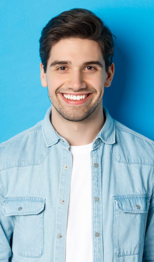 Close-up of young successful man smiling at camera, standing in casual outfit against blue background.